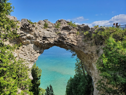 A natural stone arch overlooks a serene blue lake, surrounded by lush greenery and a clear sky.