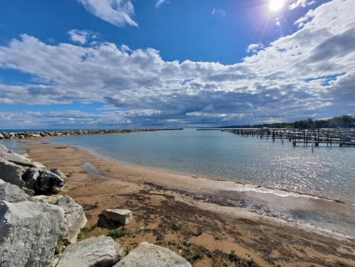 A serene beach scene with sandy shore, calm water, and cloudy blue sky, featuring distant docks and rocky formations.