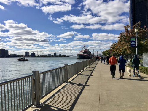 A scenic waterfront path with people walking, a boat on the water, and a clear blue sky with clouds.