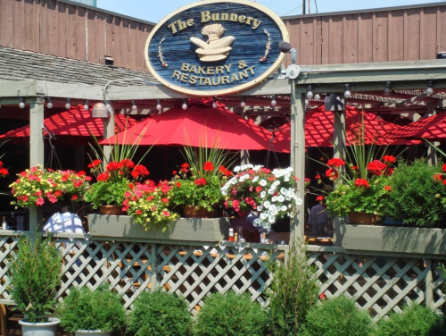 Exterior of The Bunnery Bakery & Restaurant, featuring red umbrellas, flower baskets, and a wooden sign.
