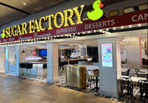 Brightly lit entrance of Sugar Factory restaurant featuring a bar, seating, and colorful signage.