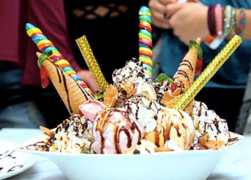 A large ice cream sundae topped with whipped cream, candies, and colorful straws, served in a white bowl.