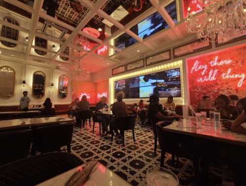 A stylish restaurant interior with chandeliers, neon signs, and patrons dining at tables.