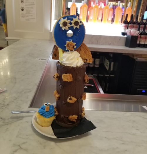 A tall chocolate dessert topped with whipped cream, a donut, and cookie pieces, alongside a small blue cupcake.