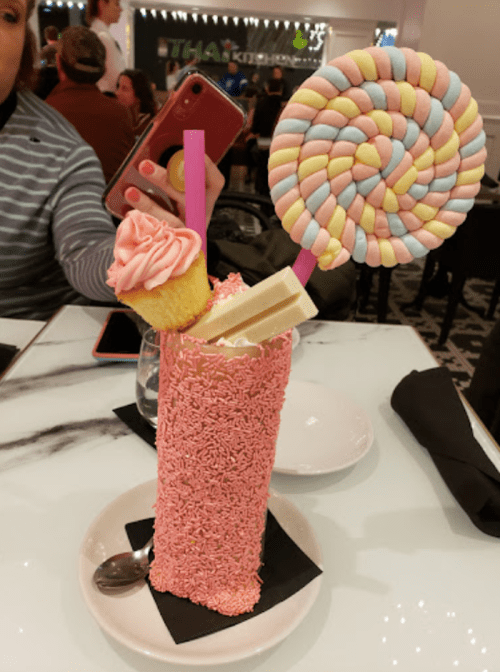 A colorful dessert with a pink sprinkle-covered cup, topped with a cupcake, lollipop, and a pink straw.