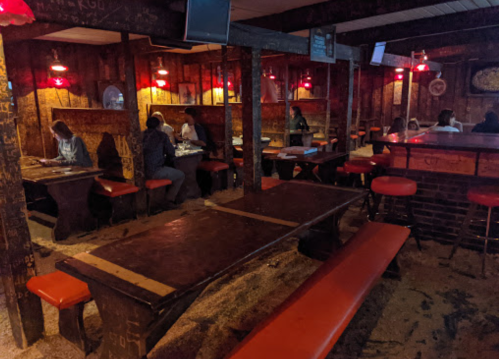A dimly lit bar with wooden tables and benches, featuring red lights and patrons seated at tables.