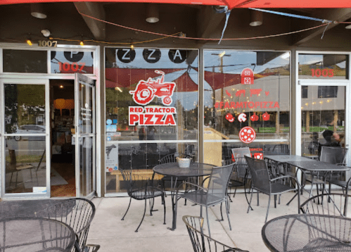 Exterior of Red Tractor Pizza with outdoor seating and a bright red awning. Large windows display the restaurant's logo.