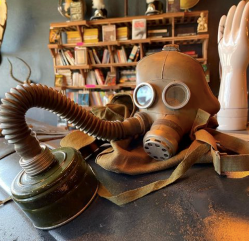 A vintage gas mask with a green canister, resting on a table near a bookshelf and a decorative hand.