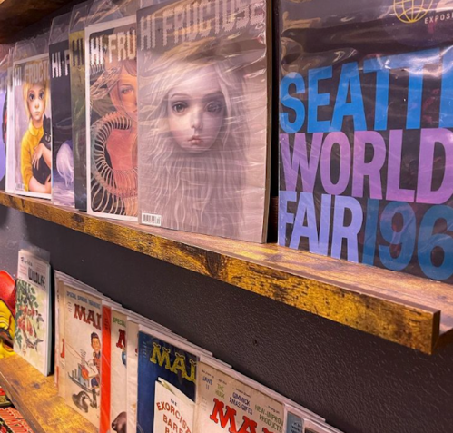 A wooden shelf displaying various magazines, including "Hi Fructose" and "Seattle World Fair 1962."