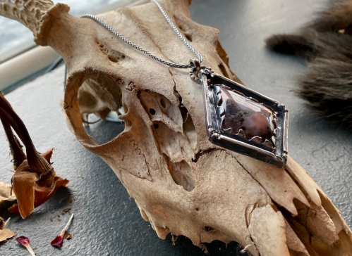 A silver necklace with a large stone pendant rests on a decorative animal skull, surrounded by dried flowers.
