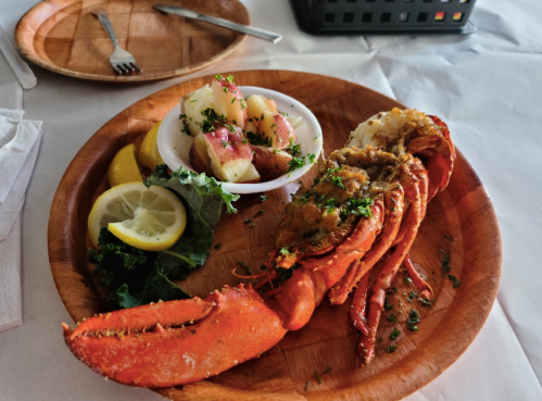 A wooden plate with a lobster tail, stuffed lobster, lemon slices, and a side of potato salad garnished with herbs.