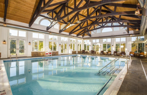 Indoor swimming pool with large windows, wooden beams, and lounge chairs around the water. Bright and inviting atmosphere.