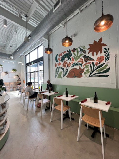 A modern restaurant interior with plants, a mural, and patrons seated at white tables with wooden chairs.