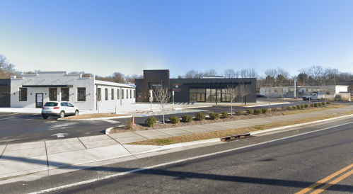 A street view of two modern buildings, one with a dark facade and the other with a light exterior, along a paved road.