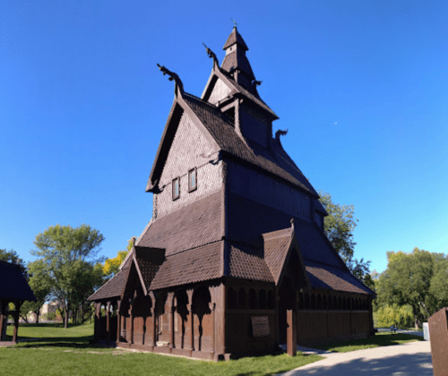 A tall, wooden church with a steep roof and decorative carvings, surrounded by trees and a clear blue sky.