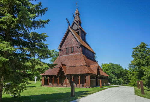 A wooden church with a steep roof, surrounded by trees and a clear blue sky. A pathway leads to the entrance.