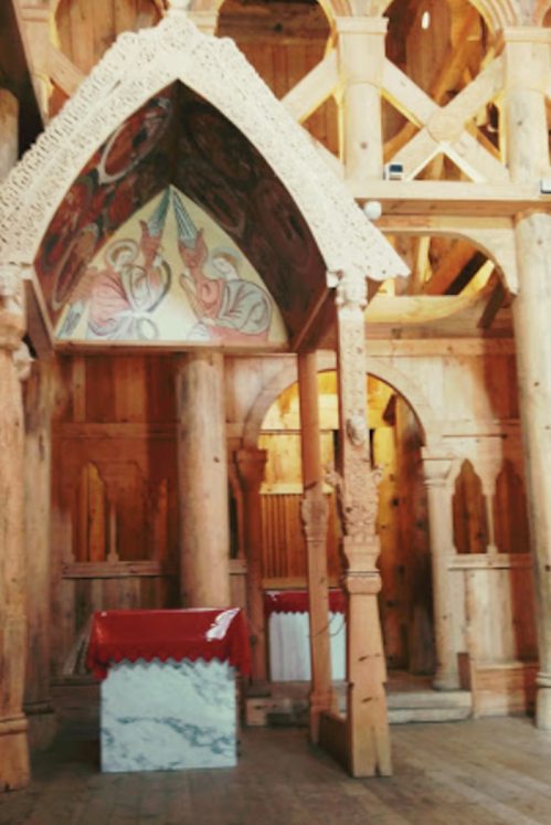 Interior of a wooden structure featuring ornate carvings, a painted ceiling, and a red-draped altar.