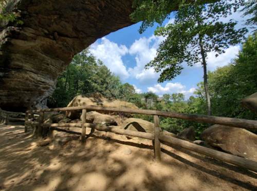 A natural stone archway surrounded by trees and a clear blue sky, with a sandy path and wooden fence in the foreground.