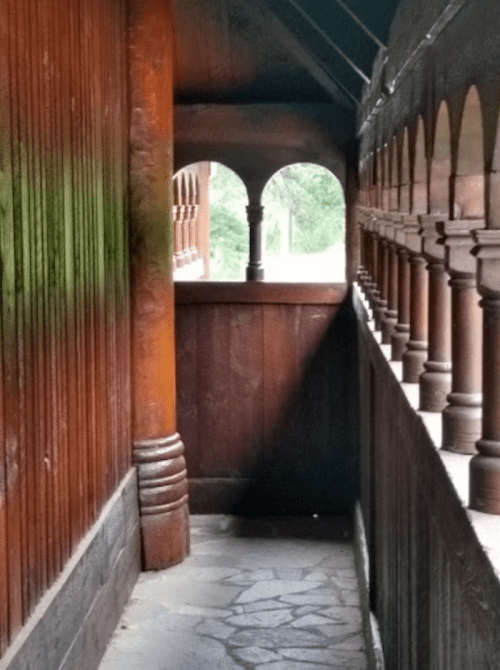 Narrow corridor with wooden walls and arches, leading to a bright outdoor view.