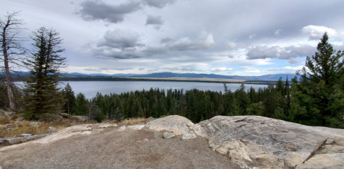A panoramic view of a lake surrounded by trees and mountains under a cloudy sky.