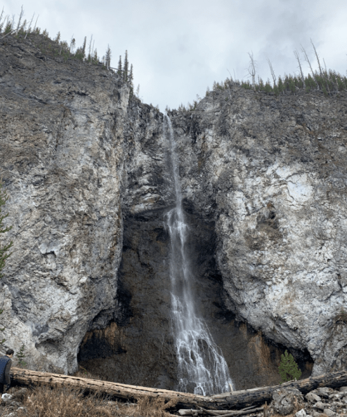 A tall waterfall cascading down a rocky cliff, surrounded by sparse trees and a cloudy sky.