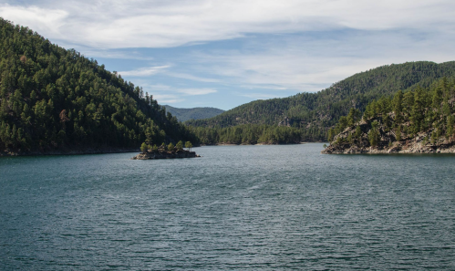 A serene lake surrounded by lush green mountains under a partly cloudy sky, with a small island in the center.