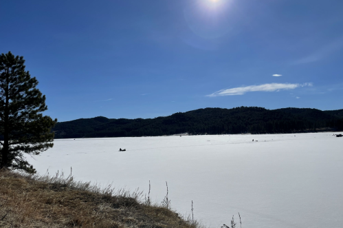 A snowy landscape with a frozen lake, surrounded by mountains and a clear blue sky, with a few people in the distance.