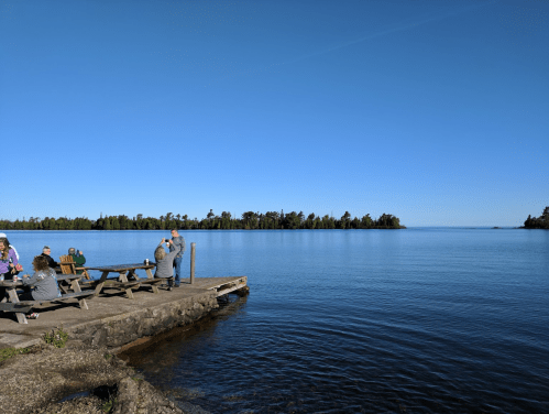 A serene lakeside scene with people sitting on a dock, surrounded by calm water and trees under a clear blue sky.