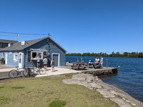 A blue building by the water with people gathered outside, bicycles nearby, and a clear blue sky above.