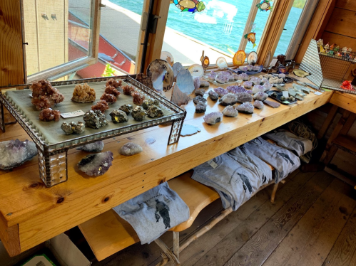 A wooden table displays various colorful stones and crystals, with T-shirts underneath and a scenic view outside.