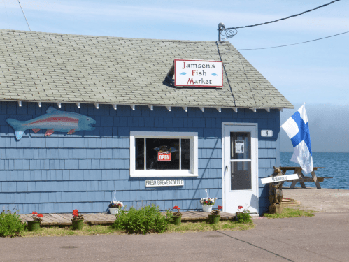 A blue fish market building with a sign reading "Jamsen's Fish Market," flowers in front, and a view of the water.