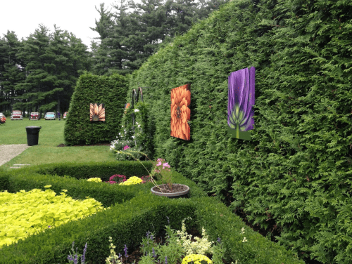 Colorful flower paintings displayed on a green hedge in a garden, surrounded by vibrant flower beds.
