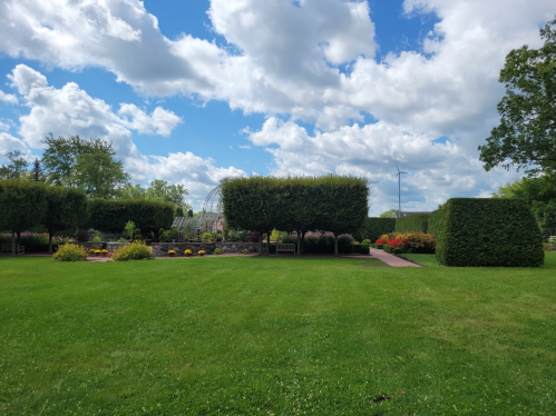 A lush green garden with neatly trimmed hedges, colorful flowers, and a blue sky dotted with clouds.