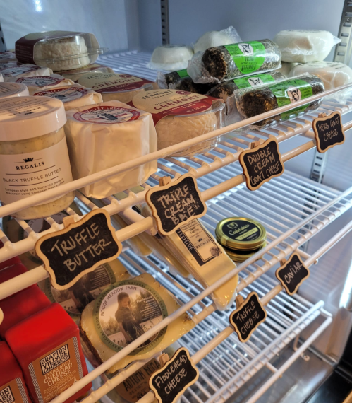A refrigerator shelf filled with various cheeses and spreads, labeled with small chalkboard tags.
