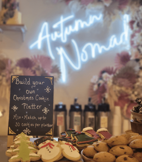 A festive display of cookies with a sign inviting customers to build their own Christmas cookie platter. Neon "Autumn Nomad" sign in background.