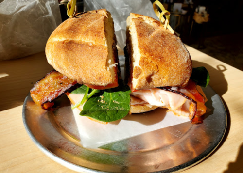 A toasted sandwich with turkey, bacon, and spinach, served on a silver plate with a sunny background.