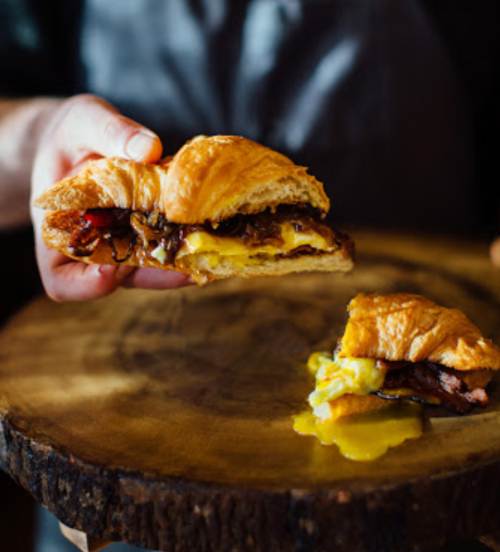A hand holding a croissant sandwich filled with bacon and egg, with a bite taken out of a second sandwich on a wooden plate.