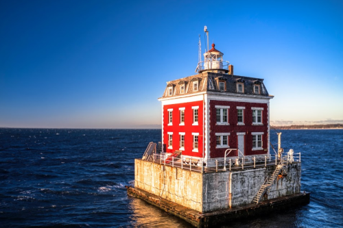 A red lighthouse stands on a rocky platform in the ocean, with clear blue skies and calm waters surrounding it.
