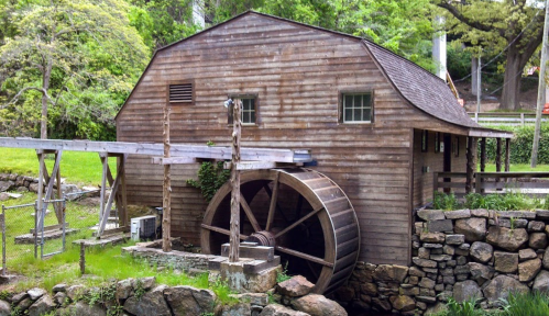 A rustic wooden mill with a large water wheel, surrounded by greenery and stone walls.