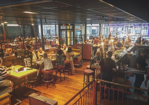A bustling restaurant interior with patrons dining at tables and a bar, featuring a lively atmosphere and wooden floors.
