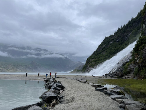 A group of hikers stands on a sandy path near a waterfall, surrounded by mountains and misty clouds.