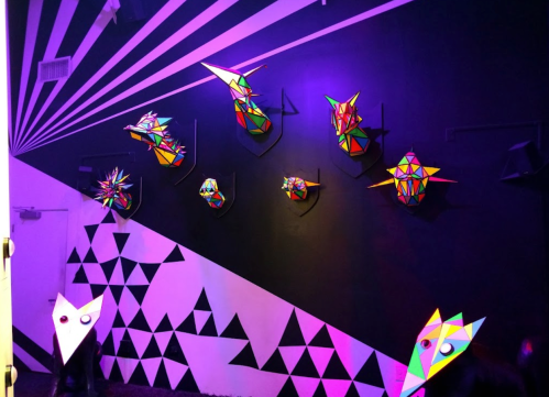 Colorful geometric sculptures mounted on a black wall, with vibrant lighting creating a dynamic art display.