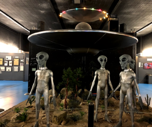 Three alien figures stand in front of a UFO display in a museum, surrounded by desert-like scenery and exhibits.