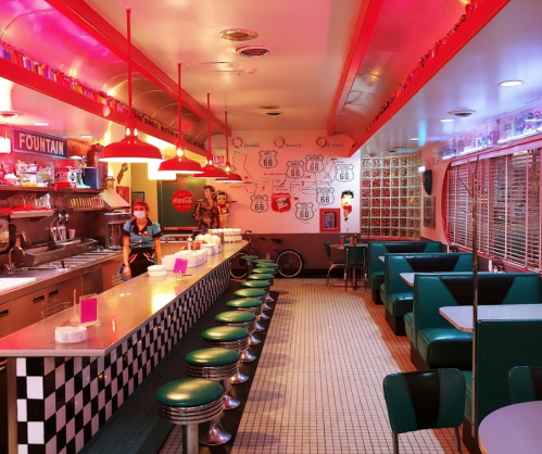 A retro diner interior with checkered floors, teal booths, and a long counter, featuring red lighting and vintage decor.