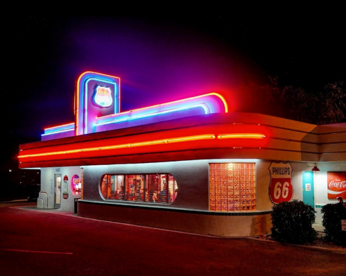 A retro diner at night, illuminated by colorful neon lights, featuring a Phillips 66 sign and large windows.
