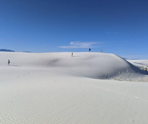 People walking on white sand dunes under a clear blue sky, with gentle hills and footprints in the sand.