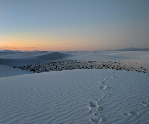 A serene desert landscape at sunset, featuring white sand dunes and footprints leading into the distance.