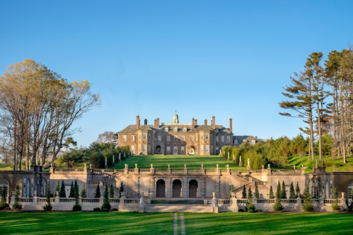 A grand mansion sits atop a lush green lawn, surrounded by trees and elegant landscaping under a clear blue sky.