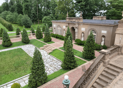 A landscaped garden featuring neatly trimmed trees, stone pathways, and a classical building with statues.