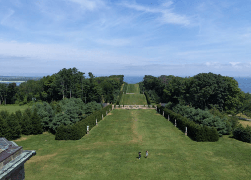 Aerial view of a lush green landscape with a long pathway, trees, and two people walking in the distance.